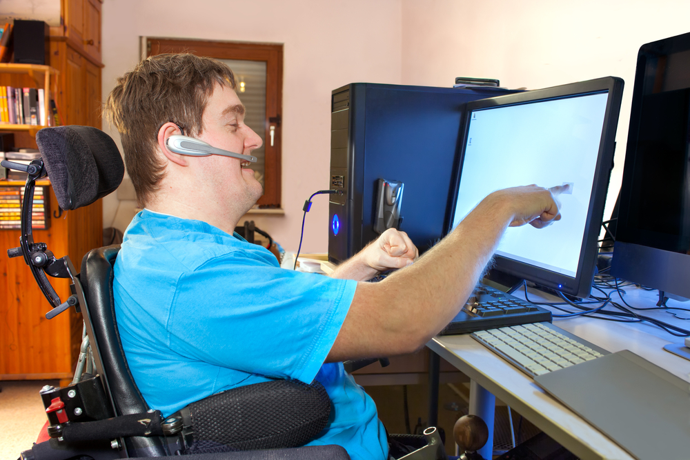 Disabled voters rights_Young, blonde, white man in blue t-shirt sits in wheelchair wearing headset and reaching to touch computer screen