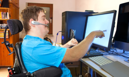 Disabled voters rights_Young, blonde, white man in blue t-shirt sits in wheelchair wearing headset and reaching to touch computer screen