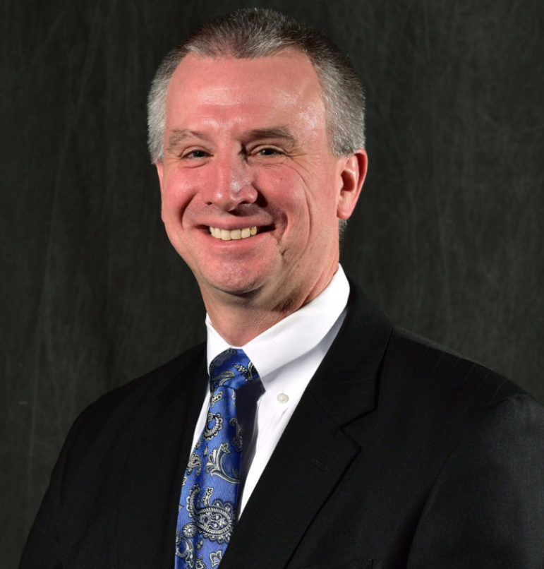 School triage training: Headshot of man with gray hair in black suit, white shirt & blue tie smiling into camera.