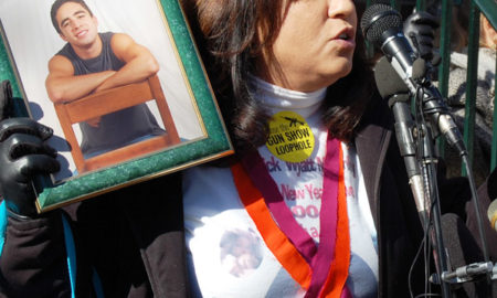 Gun shooting statistics: Woman speaking into microphones with dark hair wearing white top with black jacket and orange and purple long ribbons around her neck stands holding a green-framed photo of young man