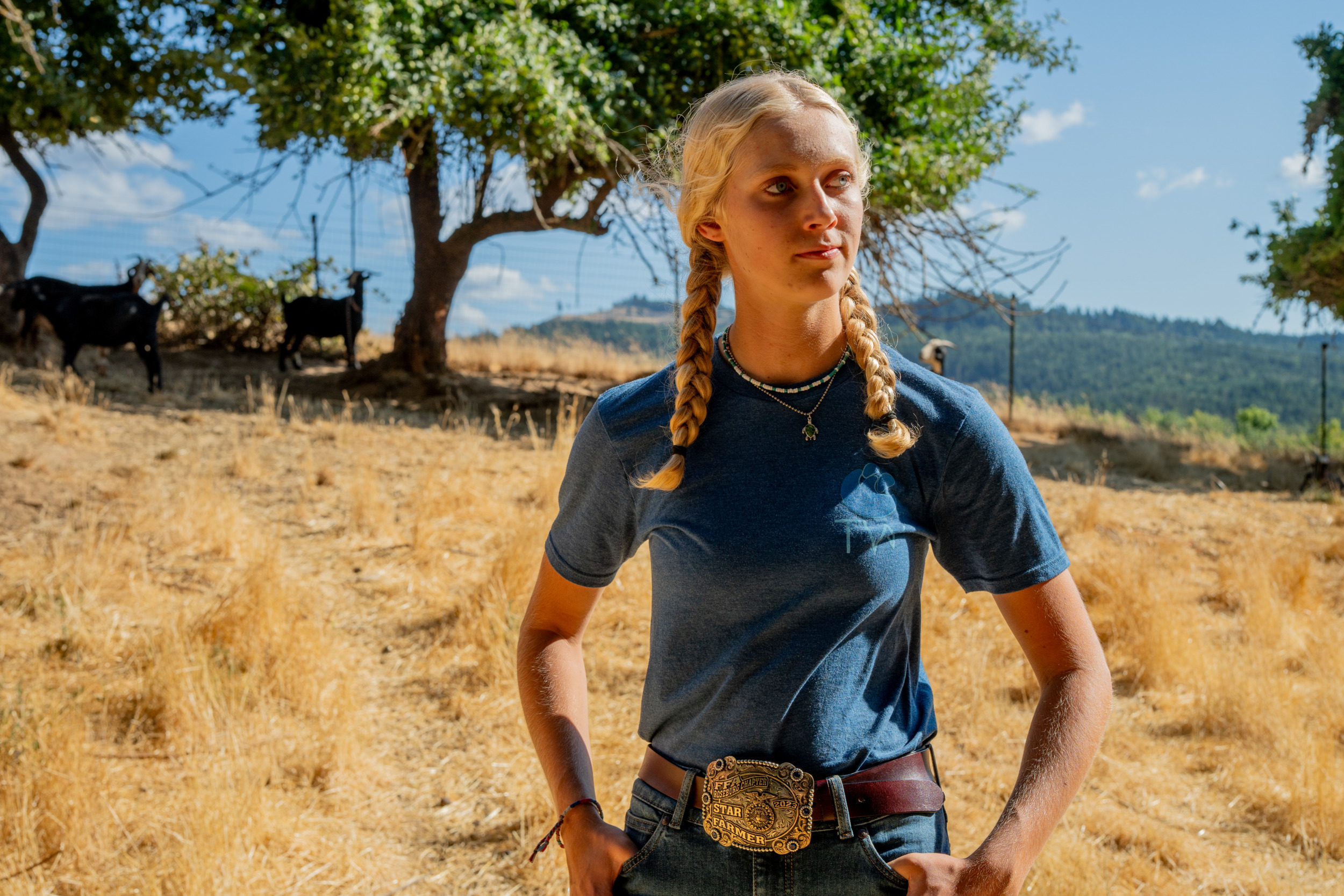 Goats - A person with two blonde braids wearing a blue t-shirt, jeans and a large belt buckle