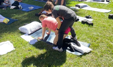 adaptive yoga: A partially paralyzed teen girl, wearing an orange shirt and shorts, gets help from an adaptive yoga instruction who is supporting and hold up the girl's torso, while the girl is on her hands and knees on a yoga mat.