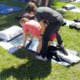 adaptive yoga: A partially paralyzed teen girl, wearing an orange shirt and shorts, gets help from an adaptive yoga instruction who is supporting and hold up the girl's torso, while the girl is on her hands and knees on a yoga mat.