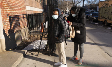 Afterschool programs virus outbreak: Black masked woman with dark hair wearing back coat andgrey leggings looking into dayback worn by black masked preteen wearing black coat with hood and tan pants, both standng on sidewalk outside brick building.