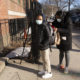 Afterschool programs virus outbreak: Black masked woman with dark hair wearing back coat andgrey leggings looking into dayback worn by black masked preteen wearing black coat with hood and tan pants, both standng on sidewalk outside brick building.