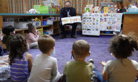 Build Back Better Child Tax Credit: Young children sit in circle on purple carpet listening to adult man in suit showing them posters