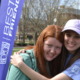 Disabled friendship program: Standing beside a blue "Best Buddies" banner, a dark-haired young woman in a white baseball cap and blue shirt and a red-headed young woman in a green sweatshirt hoodie hug each other.