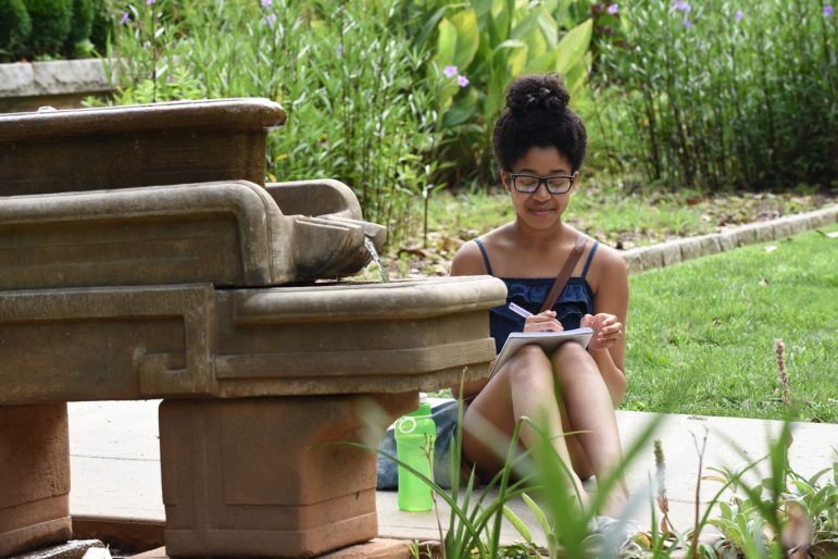 Summer Learning in OST: Young woman in shorts and tank top sitting amidst park greenery writing on notebook in her lap.