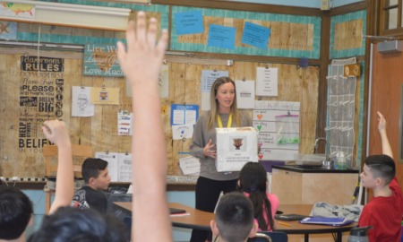 How 2 teachers use AI behind scenes: female teacher with long hair holding a white box while talking to classroom