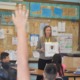 How 2 teachers use AI behind scenes: female teacher with long hair holding a white box while talking to classroom