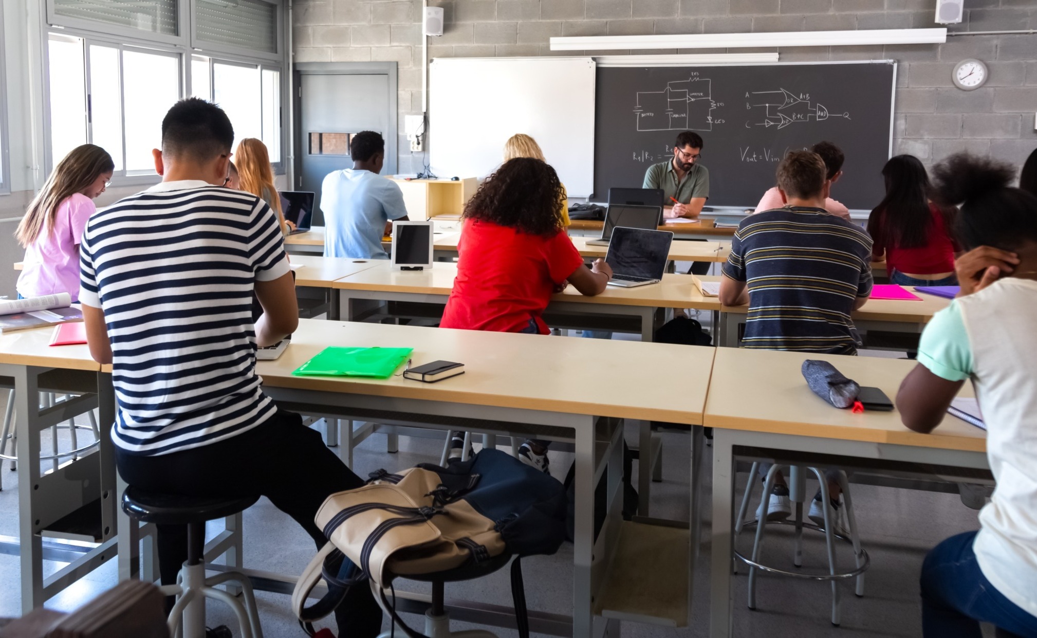 Classroom disruptions: Rear view of several students at desks bent over work with teacher facing classroom standing in front of blackboard.