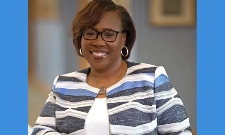 DeAngela Burns-Wallace headshot: black woman with black-rimmed glasses and colorful blazer standing with arm resting on white ledge