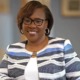 DeAngela Burns-Wallace headshot: black woman with black-rimmed glasses and colorful blazer standing with arm resting on white ledge