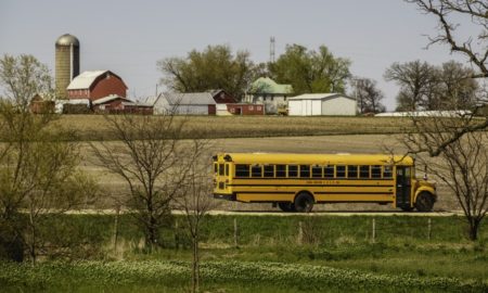 A public school bus passes by a large farmstead along a rural road on a spring day, with the edge of a county forest preserve in the foreground.