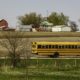 A public school bus passes by a large farmstead along a rural road on a spring day, with the edge of a county forest preserve in the foreground.