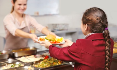 health school meals and food grants: lunch service woman hands plate of food to girl in red jacket