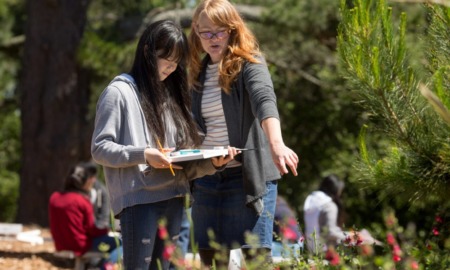 How states are using ESSER funds to strengthen high-quality OST learning: teacher with red hair teaching young Asian girl student outdoors by pointing at flowers