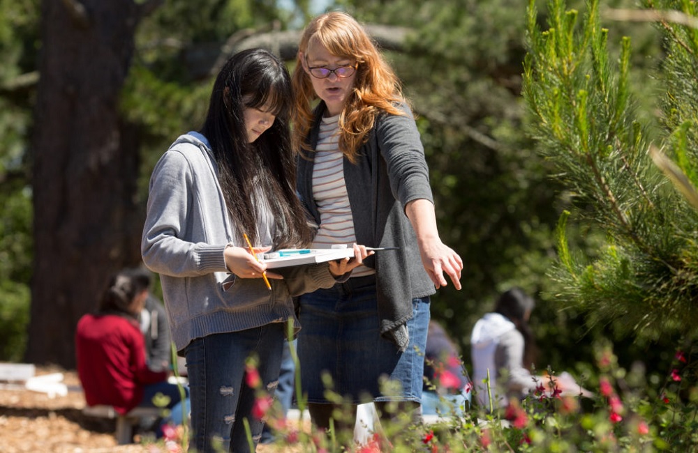 How states are using ESSER funds to strengthen high-quality OST learning: teacher with red hair teaching young Asian girl student outdoors by pointing at flowers