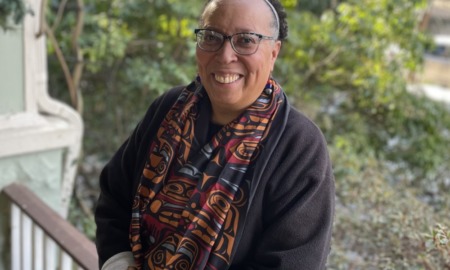 Karen Pittman opinion: Black woman with dark-frame glasses an hair pulled back stands leaning against wood porch railing wearing dark jacket and colorful scarf with greenery in the background.