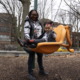 Childcare: Black woman with dark hair and black-framed glasses pushes toddler in bright yellow s playground swing