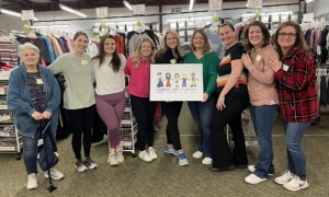 Foster care foundation: Nine women stand posing in a line smiling at the camera. Behind them are racks of hanging clothing. The two women in the middle are holding a sign featuring a graphic of children that reads "Foster Care Support www.fostercares.org"