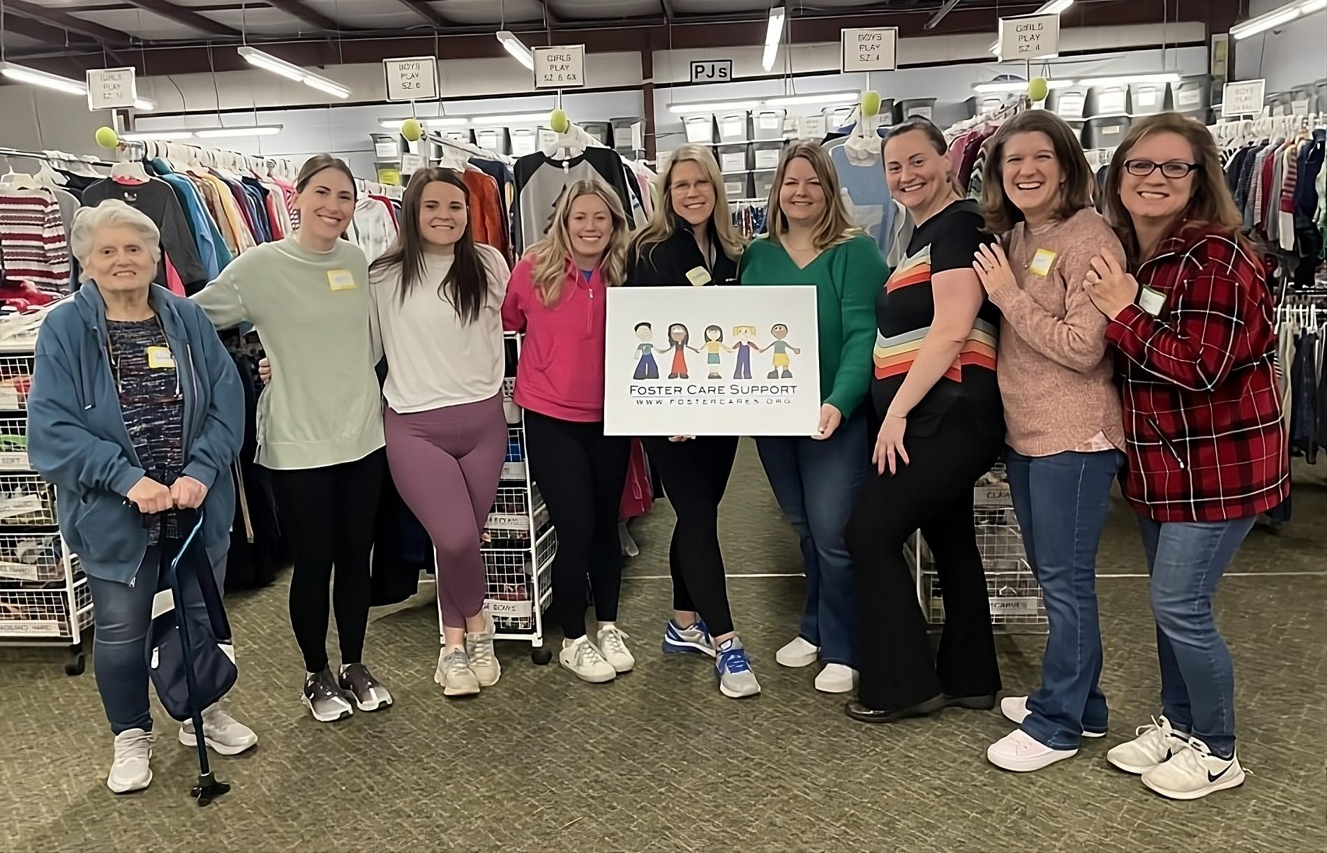 Foster care foundation: Nine women stand posing in a line smiling at the camera. Behind them are racks of hanging clothing. The two women in the middle are holding a sign featuring a graphic of children that reads "Foster Care Support www.fostercares.org"