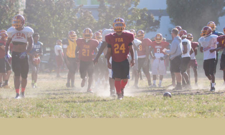 Football players at practice on a field in Harlem, dust rising from the field, players walking back towards the huddle as defense is regrouping.