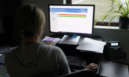 AI Algorithm child welfare assessment: ehouette of back of adult sitting at desk in front of brightly lit computer screen in dimly room.