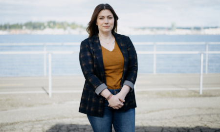 Youth homelessness Washington state: Woman with long dark hair in black jacket, copper top and blue jeans, stands on beach.