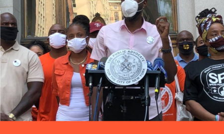 gun violence: Man in short-sleeved pink shirt, mask speaks at outdoor podium surrounded by crowd wearing masks