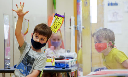 School attendance: 3 young children, one with habd raised, wearing masks sit at desks behind plexiglass dividers in frnt of a wall with colorful posters