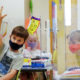 School attendance: 3 young children, one with habd raised, wearing masks sit at desks behind plexiglass dividers in frnt of a wall with colorful posters