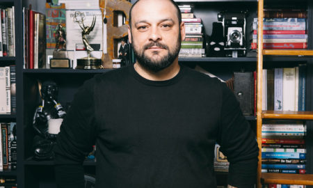 Extremism: Balding man with dark hair, beard, mustache, black top stands in front of bookshelves