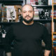 Extremism: Balding man with dark hair, beard, mustache, black top stands in front of bookshelves