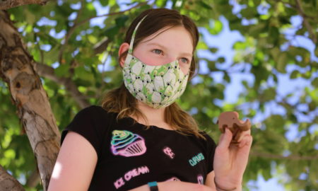 Young girl in black t-shirt wearing a mask, holds up small brown elephant sculpture in left hand, standing in front of leafy green tree