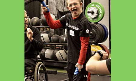 Disabled youth fitness: young man in black and red gym clothes holds workout rope standing in frong of a free weights set and next to another person in a wheelchair in a gym.