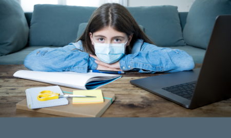 NY mental health school budget cuts: Ypung, dark-haired girl in light blue shirt wearing mask, sits at desk with head on arms