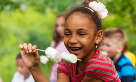 Summer Program inequities: Yooung black girl with air pulled back weaing red t-shirt, smiling holding marshmallows on a stick