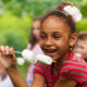 Summer Program inequities: Yooung black girl with air pulled back weaing red t-shirt, smiling holding marshmallows on a stick