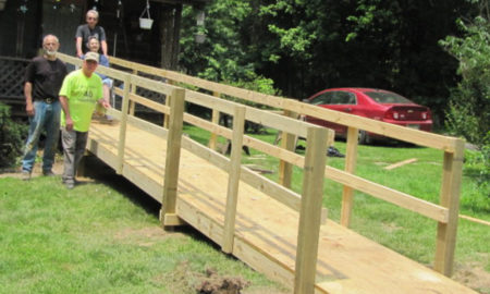 Caregiver volunteers: 3 men stand with elderly woman in wheelchair at top of new wooden handicap ramp going up to her porch across her lawn