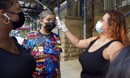 Vulnerable Students: Woman with long, curly red hair wearing mask and block tank top takes temperature of teen boy with buzz cut wearing mask and colorful superhero t-shirt standing next to another female adult wearing mask.