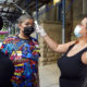 Vulnerable Students: Woman with long, curly red hair wearing mask and block tank top takes temperature of teen boy with buzz cut wearing mask and colorful superhero t-shirt standing next to another female adult wearing mask.