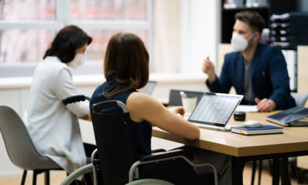 Disabled workers workplace: Three people sit around table with laptops - one person in a wheelchair - wearing face mask