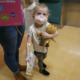 COVID and children: White, blonde toddler child wearing a pink facemask and holding a tan teddy bear stands ho.lding the hand of female adult in blue jeans and red t-shirt.