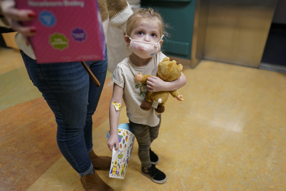 COVID and children: White, blonde toddler child wearing a pink facemask and holding a tan teddy bear stands ho.lding the hand of female adult in blue jeans and red t-shirt.