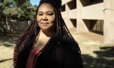 New Mexico Sex Trafficking: Middle-aged woman with long dark hair wearing black jacket with red blouse stands outside in frone of a beige stucco building and trees.