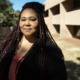 New Mexico Sex Trafficking: Middle-aged woman with long dark hair wearing black jacket with red blouse stands outside in frone of a beige stucco building and trees.