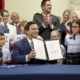 Don't Say Gay Florida: Smiling white man in blue suit sits at table holding up 2-page ocument in a display folder syrrounded by smiling white adults and children