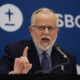 Southern Baptist Offender Man with grey hair and beard wearing dark suit, gestures with right hand while speaking into microphone at podium standing in front of blue SBC banner