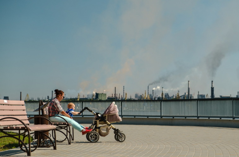 Childhood asthma: Woman sits on a park bench, while baby is in stroller, with industrial smokestacks in the background.